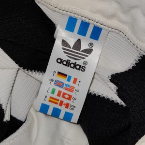 May include: Close-up of an Adidas clothing tag. The tag is white with the Adidas logo and text in black. The tag also features blue stripes and flags representing different countries. The background is white and black.