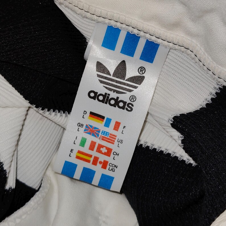May include: Close-up of an Adidas clothing tag. The tag is white with the Adidas logo and text in black. The tag also features blue stripes and flags representing different countries. The background is white and black.