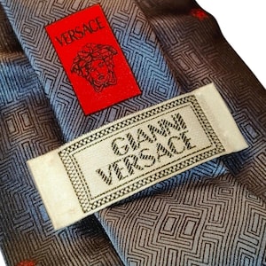 Peut inclure: Gros plan sur une cravate Versace avec un motif g&eacute;om&eacute;trique dans des tons de gris et de bleu. Une &eacute;tiquette rouge avec le logo Versace est visible, ainsi qu'une &eacute;tiquette blanche avec l'inscription "GIANNI VERSACE".