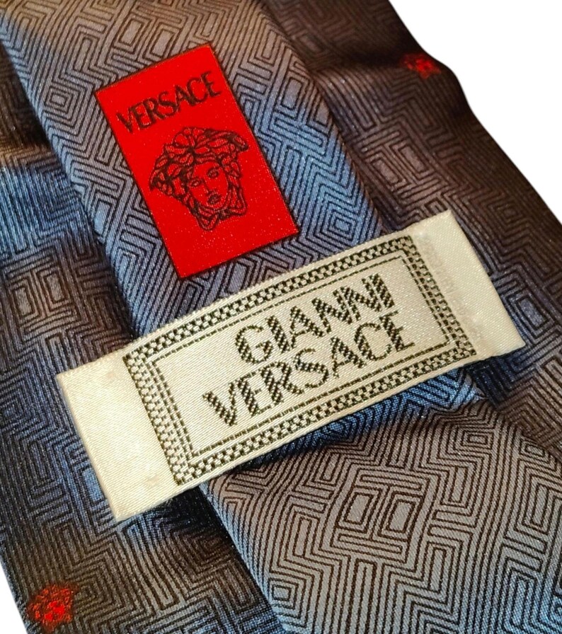 Peut inclure: Gros plan sur une cravate Versace avec un motif g&eacute;om&eacute;trique dans des tons de gris et de bleu. Une &eacute;tiquette rouge avec le logo Versace est visible, ainsi qu'une &eacute;tiquette blanche avec l'inscription "GIANNI VERSACE".