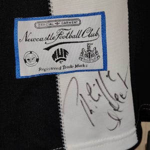 May include: Close-up of a black and white striped jersey with a Newcastle Football Club label. The label is white with blue trim and features the club's logo and text. A signature is visible on the jersey.