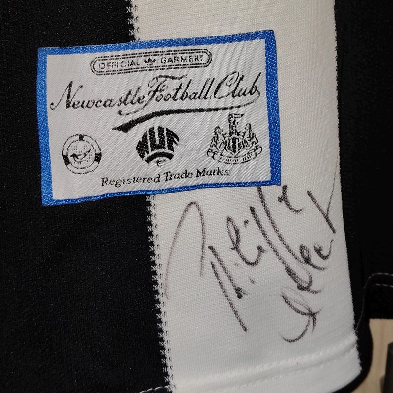 May include: Close-up of a black and white striped jersey with a Newcastle Football Club label. The label is white with blue trim and features the club's logo and text. A signature is visible on the jersey.