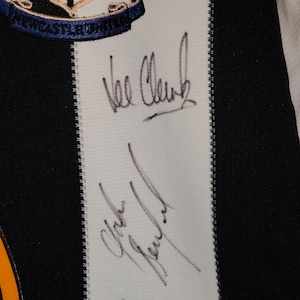 May include: Close-up of a black and white striped jersey with the Newcastle United crest at the top. The jersey features two signatures in black ink on a white stripe. The jersey has a black and white striped design.