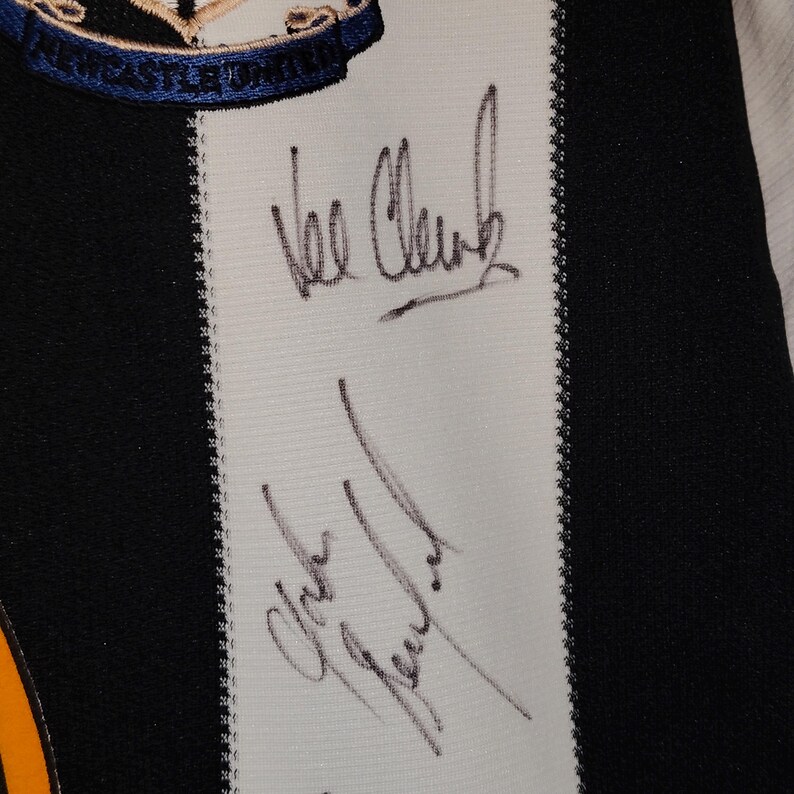 May include: Close-up of a black and white striped jersey with the Newcastle United crest at the top. The jersey features two signatures in black ink on a white stripe. The jersey has a black and white striped design.