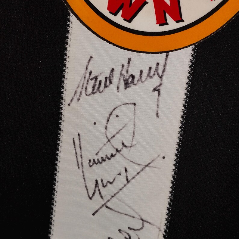 May include: Close-up of a black and white jersey with a white stripe down the center. The stripe features handwritten signatures in black ink. The top of the jersey has a circular logo with red and yellow text.