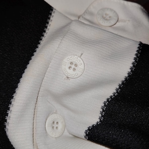 May include: Close-up of a white and black polo shirt. The shirt features white buttons with the Adidas logo. The collar and button placket are white, while the sleeves and side panels are black. The stitching is visible.