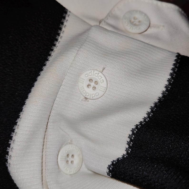 May include: Close-up of a white and black polo shirt. The shirt features white buttons with the Adidas logo. The collar and button placket are white, while the sleeves and side panels are black. The stitching is visible.