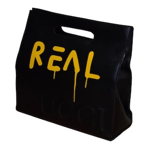 May include: Black tote bag with a cut-out handle. The word "REAL" is printed in yellow, dripping down the side. The word "GUCCI" is embossed at the bottom of the bag.