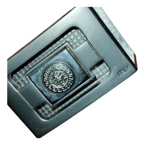 K&ouml;nnte beinhalten: Ein silberfarbener Geldclip aus Metall mit einem zentralen Quadrat, das ein erhabenes, kreisf&ouml;rmiges Emblem mit einem L&ouml;wenkopf aufweist. Der Clip hat ein geometrisches Design mit einem strukturierten Gittermuster und das Wort "ITALY" ist sichtbar.