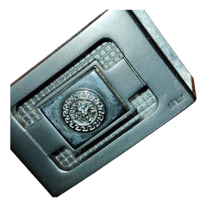 K&ouml;nnte beinhalten: Ein silberfarbener Geldclip aus Metall mit einem zentralen Quadrat, das ein erhabenes, kreisf&ouml;rmiges Emblem mit einem L&ouml;wenkopf aufweist. Der Clip hat ein geometrisches Design mit einem strukturierten Gittermuster und das Wort "ITALY" ist sichtbar.
