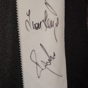 May include: Close-up of a white fabric panel with black vertical stripes on the left and right edges. Two signatures are written in black ink on the white panel. The fabric has a textured appearance.