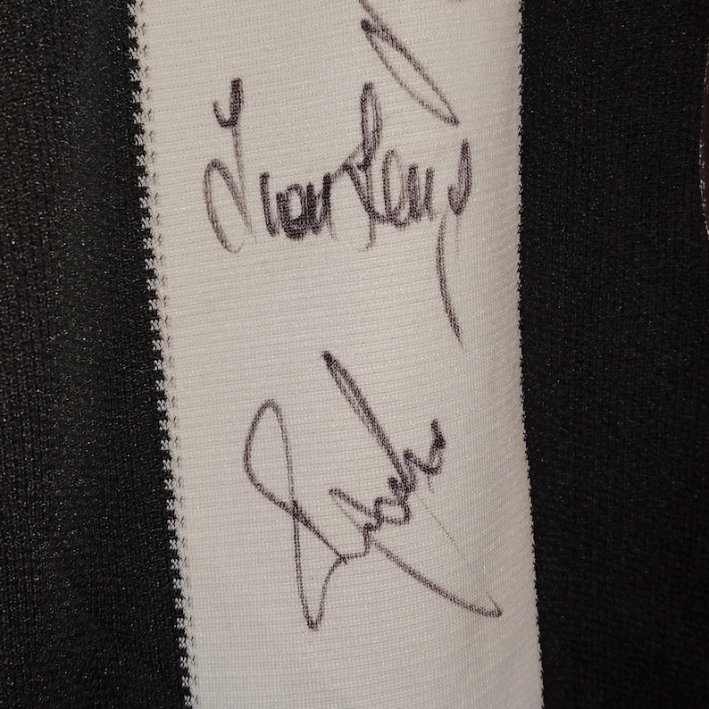 May include: Close-up of a white fabric panel with black vertical stripes on the left and right edges. Two signatures are written in black ink on the white panel. The fabric has a textured appearance.