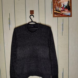 Könnte beinhalten: Ein langärmeliger Pullover mit Rundhalsausschnitt in einem dunkel anthrazitfarbenen und schwarzen melierten Strickmuster. Das Kleidungsstück weist eine gerippte Textur an Körper, Ärmeln und Saum auf, mit einem schwarzen Innenetikett am Halsausschnitt.