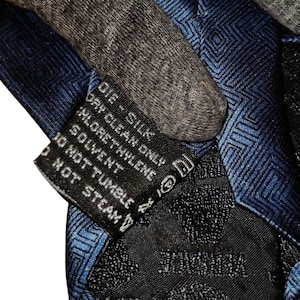 Peut inclure: Gros plan sur une cravate en soie bleue avec un motif g&eacute;om&eacute;trique. La cravate a une &eacute;tiquette noire avec des instructions d'entretien et le mot "VERSACE". La cravate est en soie.