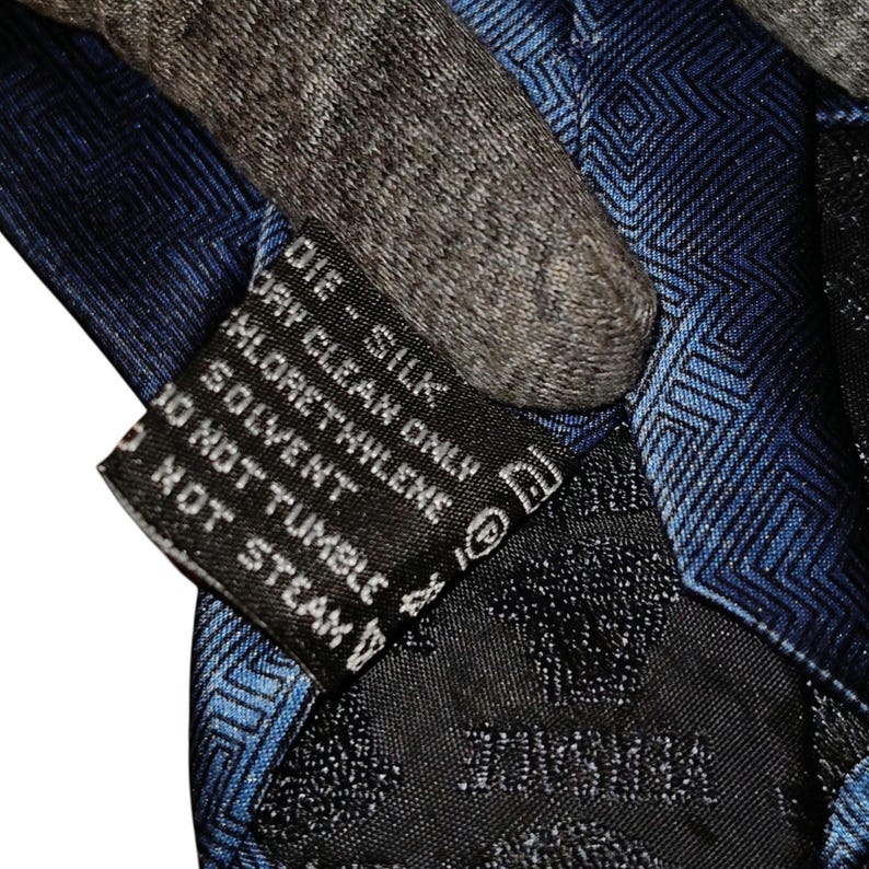 Peut inclure: Gros plan sur une cravate en soie bleue avec un motif g&eacute;om&eacute;trique. La cravate a une &eacute;tiquette noire avec des instructions d'entretien et le mot "VERSACE". La cravate est en soie.