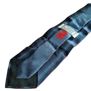 Peut inclure: Une cravate en soie bleue avec un motif g&eacute;om&eacute;trique et de petits accents rouges. La cravate porte une &eacute;tiquette avec le texte "GIANNI VERSACE" et un logo Versace rouge. La cravate est pli&eacute;e, r&eacute;v&eacute;lant l'envers avec un motif plus fonc&eacute;.