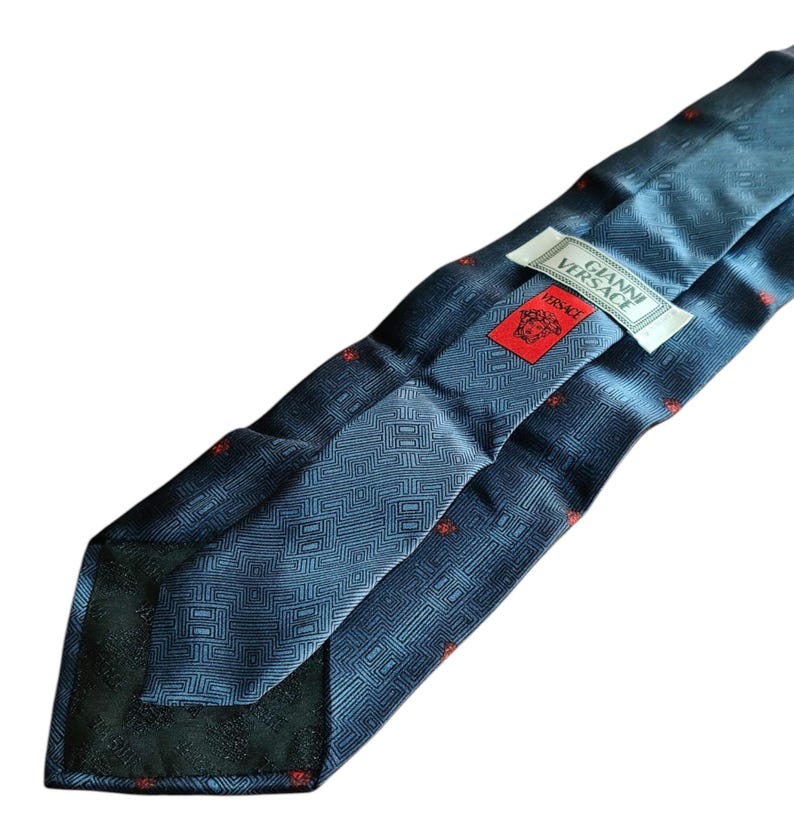 Peut inclure: Une cravate en soie bleue avec un motif g&eacute;om&eacute;trique et de petits accents rouges. La cravate porte une &eacute;tiquette avec le texte "GIANNI VERSACE" et un logo Versace rouge. La cravate est pli&eacute;e, r&eacute;v&eacute;lant l'envers avec un motif plus fonc&eacute;.