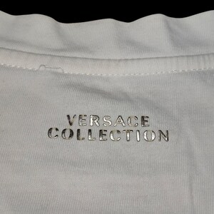 May include: A white fabric garment featuring the text VERSACE COLLECTION printed in a metallic silver font. The text is centered below the neckline, which shows a visible stitched seam along the collar area of the white textile material.