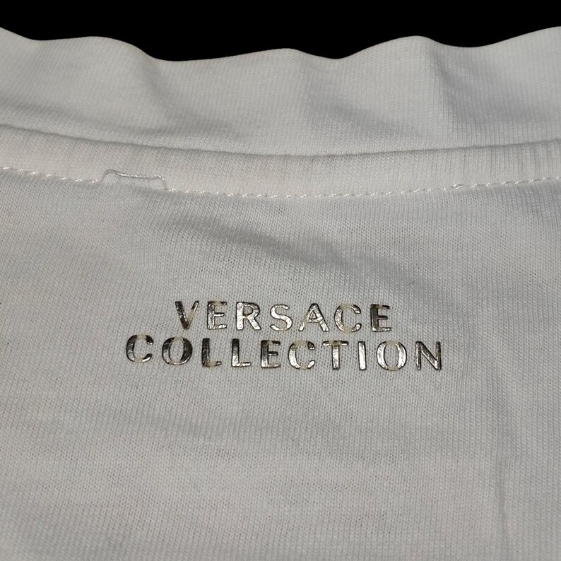 May include: A white fabric garment featuring the text VERSACE COLLECTION printed in a metallic silver font. The text is centered below the neckline, which shows a visible stitched seam along the collar area of the white textile material.