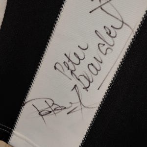 May include: Close-up of a black and white striped fabric with the handwritten signature "Peter Beardsley" in black ink. The signature is on a white stripe, with a black stripe visible.