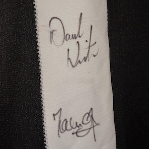 May include: Close-up of a white fabric strip with two signatures in black ink. The fabric is bordered by black and white striped material. The signatures appear to be handwritten.