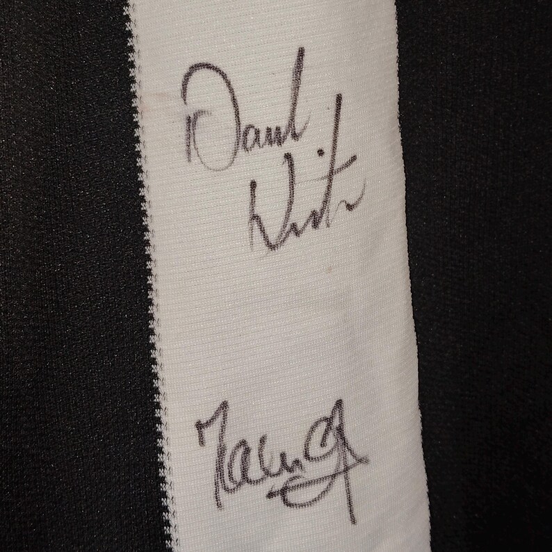 May include: Close-up of a white fabric strip with two signatures in black ink. The fabric is bordered by black and white striped material. The signatures appear to be handwritten.