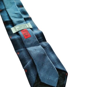 Peut inclure: Cravate en soie bleue avec un motif g&eacute;om&eacute;trique et de petits accents rouges. La cravate porte l'&eacute;tiquette "GIANNI VERSACE" et une &eacute;tiquette rouge avec le logo Versace.