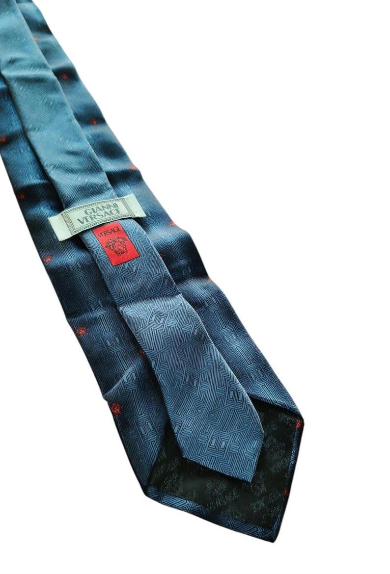 Peut inclure: Cravate en soie bleue avec un motif g&eacute;om&eacute;trique et de petits accents rouges. La cravate porte l'&eacute;tiquette "GIANNI VERSACE" et une &eacute;tiquette rouge avec le logo Versace.