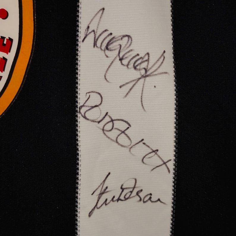 May include: Close-up of a black and white textile with three signatures in black ink. The signatures are on a white strip of fabric with a black background. The edge of a circular emblem is visible.