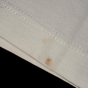 May include: A close-up view of a light cream-colored fabric featuring a double-stitched hem line running horizontally across the top. Two small, distinct brownish-orange circular stains are visible on the surface of the textile material.