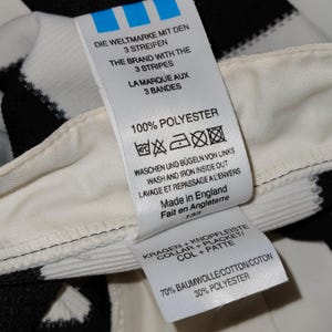 May include: Close-up of a white tag with black text, attached to a black and white striped fabric. The tag features the Adidas logo and washing instructions. The tag reads "100% Polyester" and "Made in England."