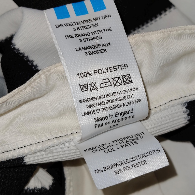 May include: Close-up of a white tag with black text, attached to a black and white striped fabric. The tag features the Adidas logo and washing instructions. The tag reads "100% Polyester" and "Made in England."