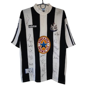 May include: Black and white striped soccer jersey with the Adidas logo and Newcastle Brown Ale logo. The jersey has multiple signatures and a crest. The jersey is hanging on a hanger.