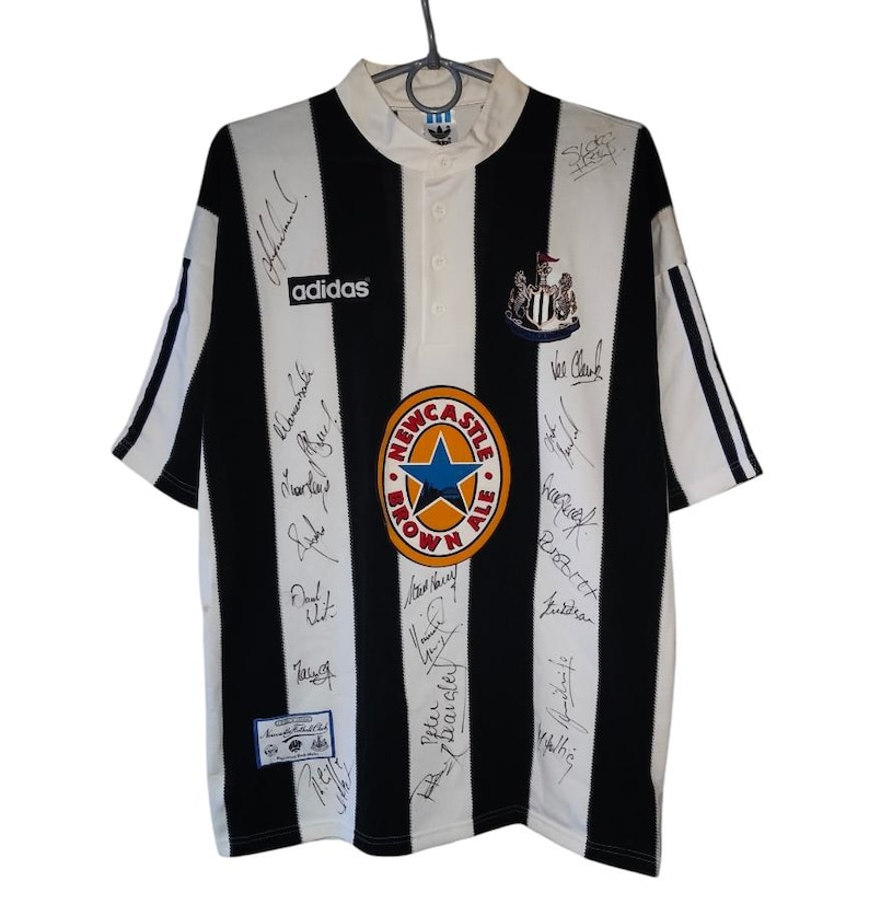 May include: Black and white striped soccer jersey with the Adidas logo and Newcastle Brown Ale logo. The jersey has multiple signatures and a crest. The jersey is hanging on a hanger.