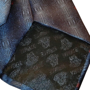 Peut inclure: Gros plan sur une cravate &agrave; motifs bleus et noirs avec un motif g&eacute;om&eacute;trique. La cravate a un dos noir avec le mot "VERSACE" imprim&eacute; dessus. Des accents rouges sont visibles dans tout le motif.