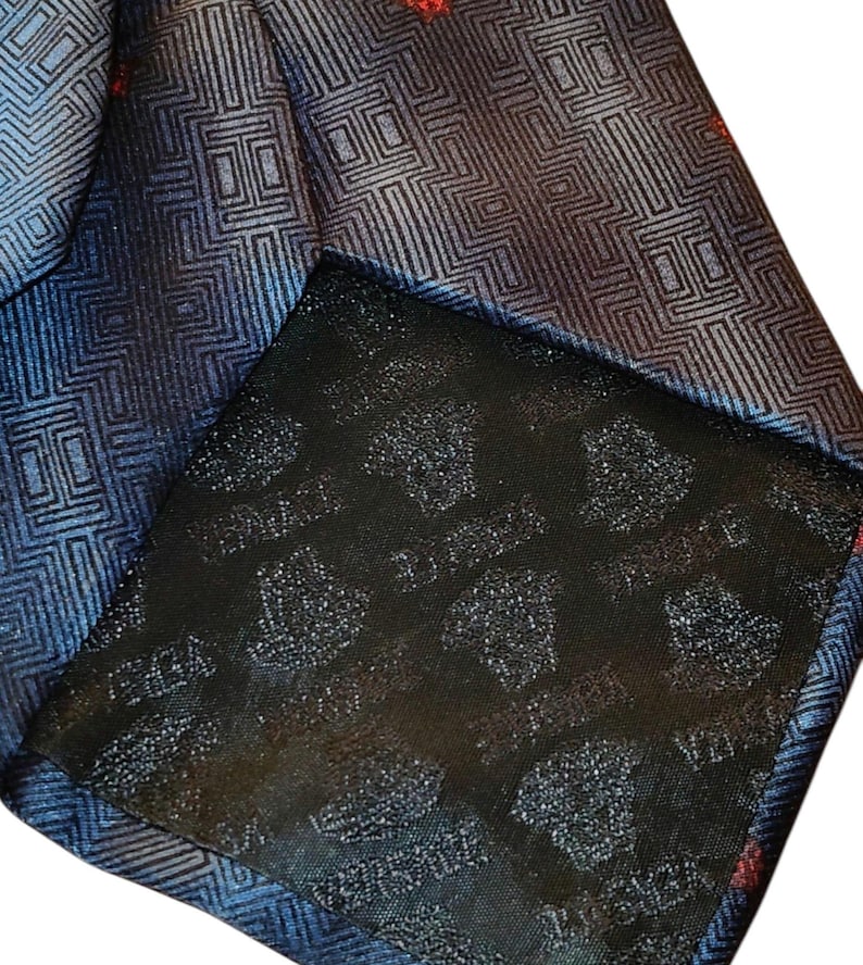 Peut inclure: Gros plan sur une cravate &agrave; motifs bleus et noirs avec un motif g&eacute;om&eacute;trique. La cravate a un dos noir avec le mot "VERSACE" imprim&eacute; dessus. Des accents rouges sont visibles dans tout le motif.