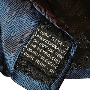 Peut inclure: Gros plan sur une cravate en soie bleue et noire avec un motif g&eacute;om&eacute;trique. L'&eacute;tiquette de la cravate indique "100% SETA - S" et d'autres instructions d'entretien. La cravate est en soie.