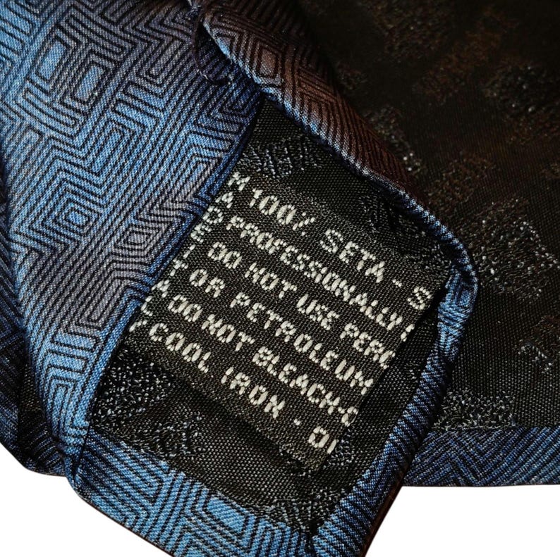 Peut inclure: Gros plan sur une cravate en soie bleue et noire avec un motif g&eacute;om&eacute;trique. L'&eacute;tiquette de la cravate indique "100% SETA - S" et d'autres instructions d'entretien. La cravate est en soie.