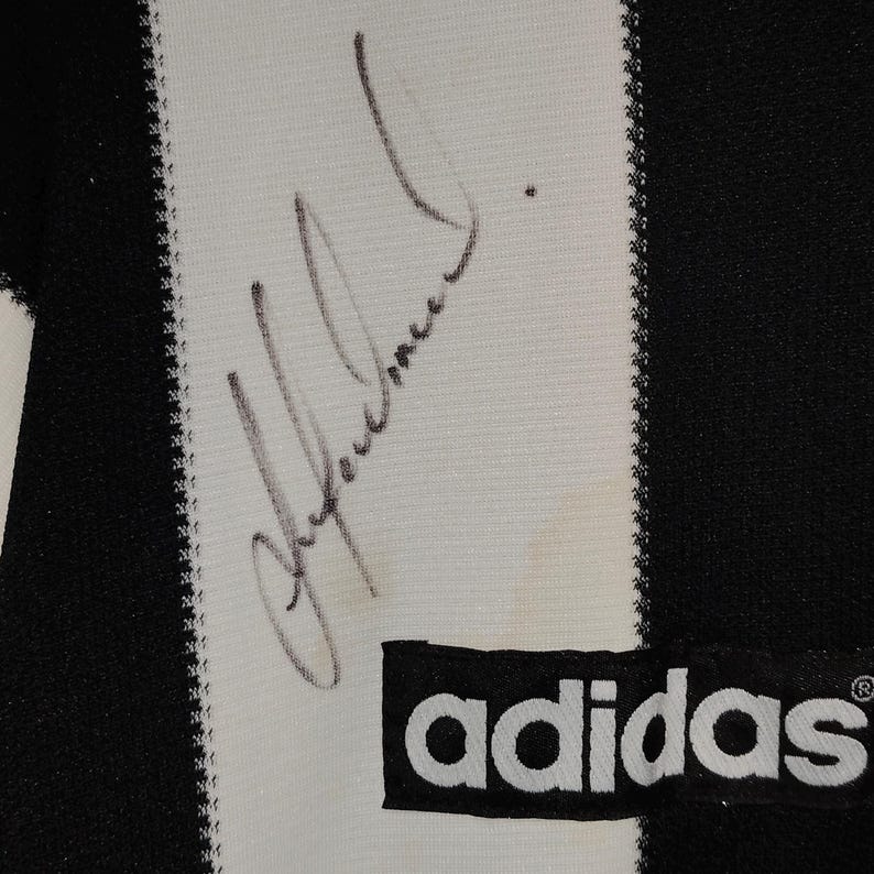 May include: Close-up of a black and white striped Adidas jersey. The jersey features a printed signature in black ink on a white stripe. The Adidas logo is visible in the lower right corner.