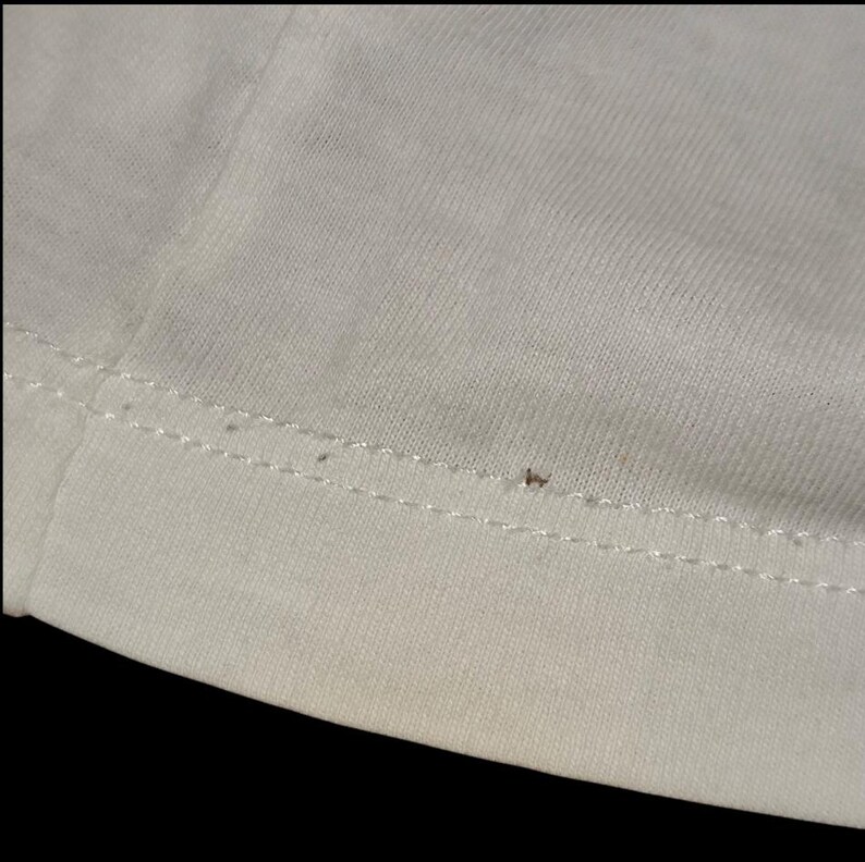 May include: A close-up view of a white fabric garment featuring a horizontal double-stitched hemline. The material displays a fine knit texture with a small, singular brown speck visible along the stitching line of the white textile surface.