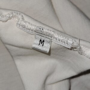 May include: A white fabric garment featuring a small, rectangular clothing tag sewn into the interior seam. The tag is white with the letter M printed in black, indicating the size. The fabric shows a visible stitched hem along the edge.
