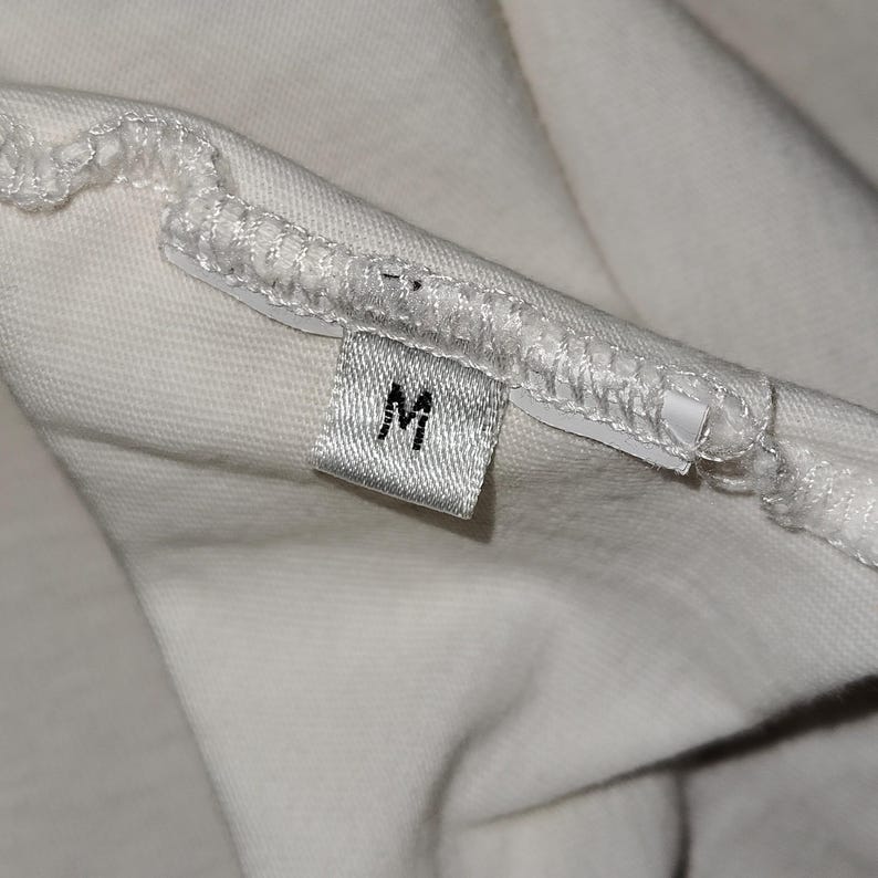 May include: A white fabric garment featuring a small, rectangular clothing tag sewn into the interior seam. The tag is white with the letter M printed in black, indicating the size. The fabric shows a visible stitched hem along the edge.