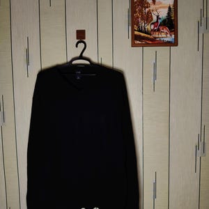 Könnte beinhalten: Ein einfarbiger schwarzer Langarmpullover mit V-Ausschnitt, der auf einem schwarzen Kunststoffbügel hängt. Das Kleidungsstück hat ein schlichtes Design mit gerippten Bündchen an Ärmeln und Saum und zeigt ein sauberes, einheitliches Erscheinungsbild.