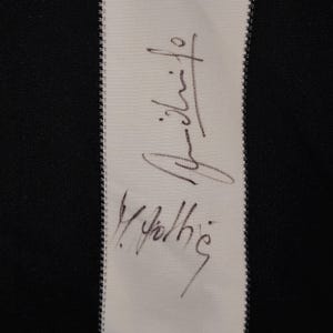 May include: Close-up of a white fabric strip with a black and white striped background. The fabric strip features a handwritten signature in black ink. The background is black and white.
