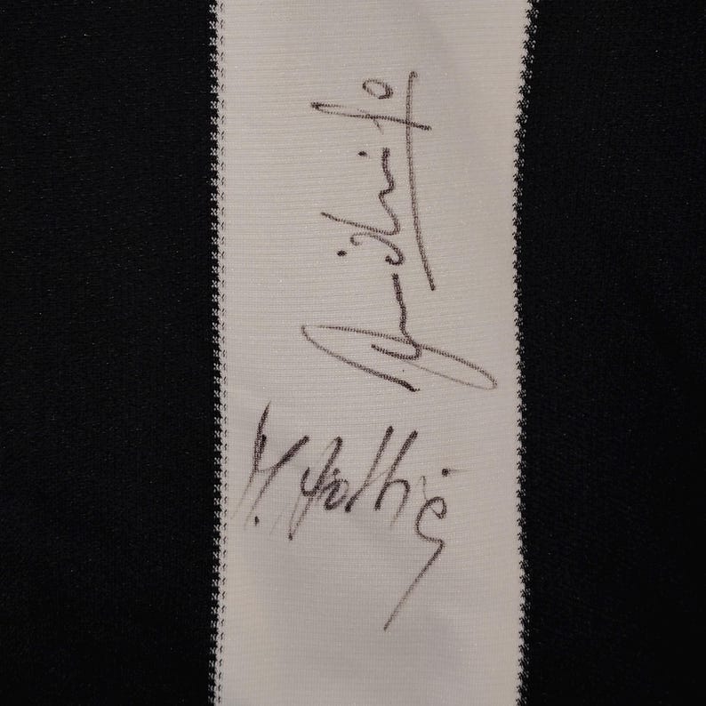 May include: Close-up of a white fabric strip with a black and white striped background. The fabric strip features a handwritten signature in black ink. The background is black and white.