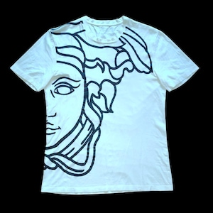 May include: A white short-sleeve t-shirt featuring a large, bold black line art graphic of a classical face with flowing hair covering the left side and extending across the upper portion of the garment, set against a plain white background.