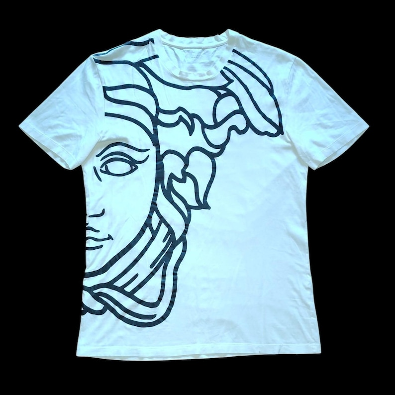 May include: A white short-sleeve t-shirt featuring a large, bold black line art graphic of a classical face with flowing hair covering the left side and extending across the upper portion of the garment, set against a plain white background.