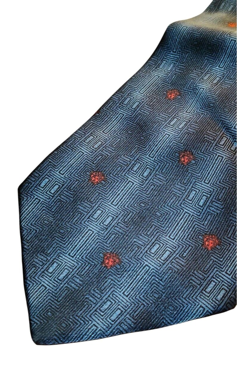Peut inclure: Une cravate bleue avec un motif g&eacute;om&eacute;trique et de petits accents rouges. La cravate pr&eacute;sente un motif r&eacute;p&eacute;titif de carr&eacute;s et de lignes, avec des accents rouges espac&eacute;s sur le tissu. La cravate est faite d'un mat&eacute;riau brillant.