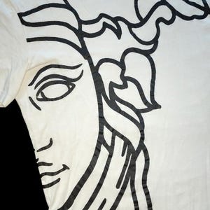 May include: A white short-sleeved t-shirt featuring a large, bold black graphic print of a stylized face with flowing hair. The design covers the front of the garment, extending from the neckline down to the hem, with clean, thick black lines.