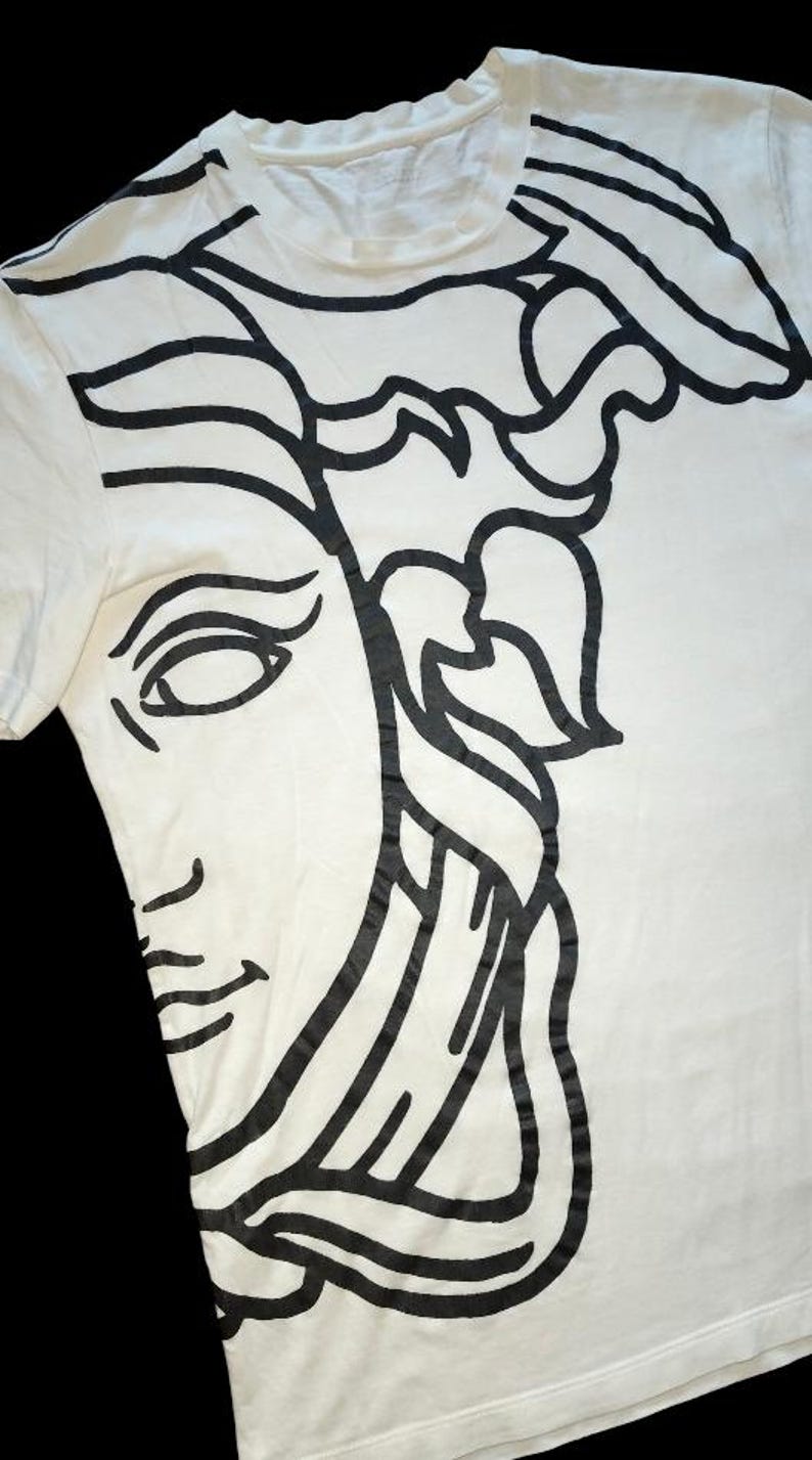 May include: A white short-sleeved t-shirt featuring a large, bold black graphic print of a stylized face with flowing hair. The design covers the front of the garment, extending from the neckline down to the hem, with clean, thick black lines.